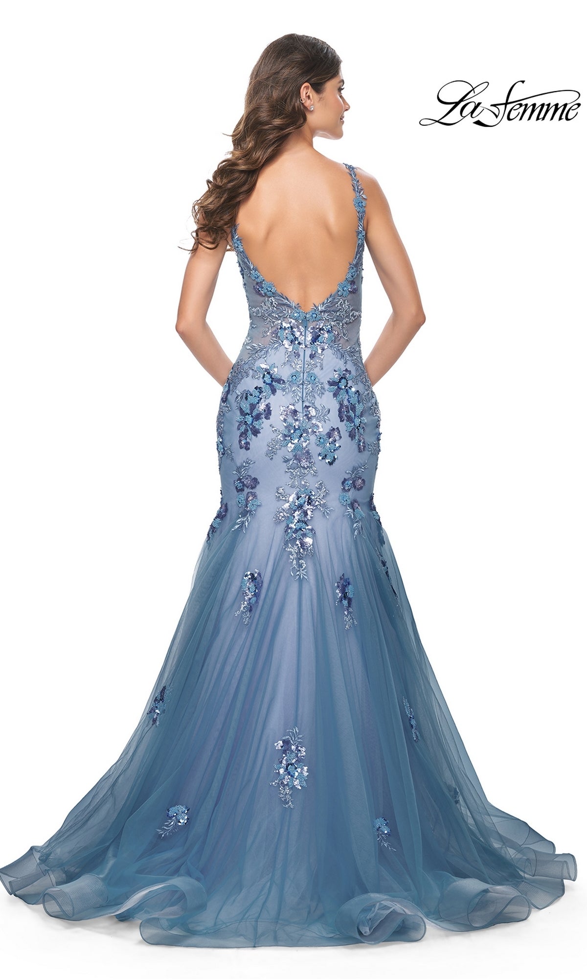 Woman in the La Femme Slate Blue Long Mermaid Prom Dress 32192, featuring a fitted silhouette, floral embellishments, and a deep V-back, standing with her back facing the camera.